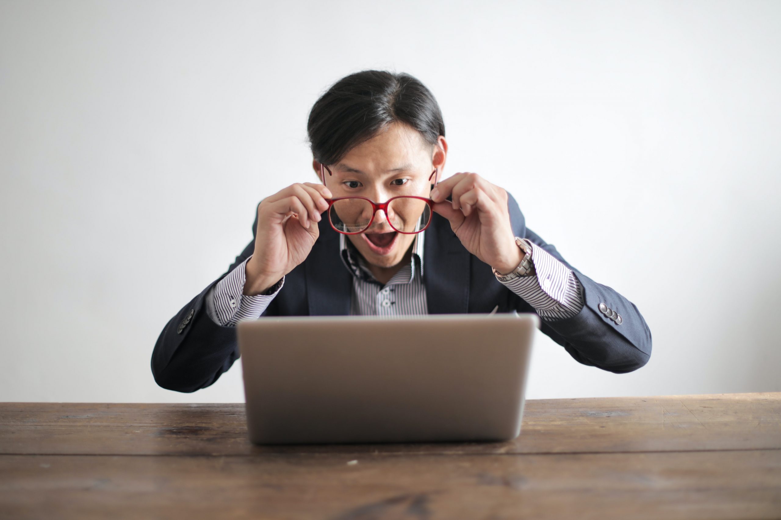 A man sitting on a desk in front of a laptop looking surprised and putting his glasses off in surprise.